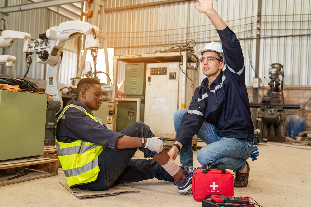 Mature foreman in workwear giving first aid to staff suffering after working injury in industrial factory, Injury at work.